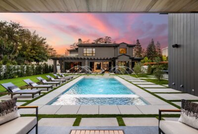 Modern luxury home with a swimming pool and sun loungers at sunset, featuring a well-manicured garden and outdoor seating area.